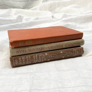 Könnte beinhalten: Ein Stapel von drei alten Büchern mit abgenutzten Einbänden. Das oberste Buch ist in einem gedämpften Orange gehalten, das mittlere Buch ist grau und das untere Buch ist braun. Auf den Buchrücken sind Texte wie "MRS OVER THE WAYS" und "BALDWIN'S READERS-SEVENTH YEAR" zu sehen.