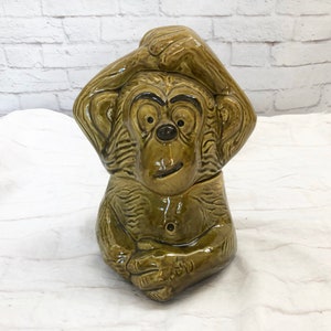 May include: An olive green ceramic monkey-shaped cookie jar. The monkey is shown with its hand on its head and a button on its chest. The jar is on a white surface, with a white brick wall in the background.