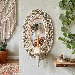May include: An oval, off-white woven wall mirror with a candle holder. The mirror has a woven frame and a candle is lit in the holder below. The wall is painted a light green, and there are plants in the background.
