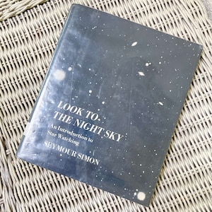 Puede incluir: Un libro titulado "Look to the Night Sky" con una cubierta gris oscuro con estrellas blancas. El libro es una introducción a la observación de estrellas de Seymour Simon. El libro está sobre una superficie de mimbre tejido.