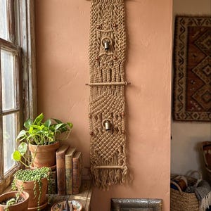 May include: A beige macrame wall hanging with wooden dowels and beads, and two small bells. The wall decor is displayed on a peach-colored wall, near a window with potted plants and books.