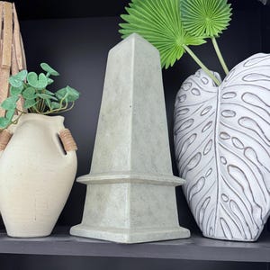 May include: A light green obelisk-shaped decorative object with a square base and a pointed top. The obelisk is displayed on a black shelf alongside a white vase with a leaf design and a cream-colored vase with a plant.