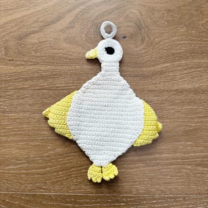 May include: A handmade crochet duckling, featuring a white body, yellow wings, and feet. The duckling has a yellow beak and a black eye. A loop is attached to the top for hanging. The craft is made with a tight stitch.