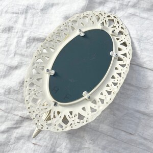May include: Ivory-colored, oval-shaped hand mirror with a decorative, openwork frame. The mirror has a dark gray reflective surface and is attached with small screws. The handle is a continuation of the frame's design.