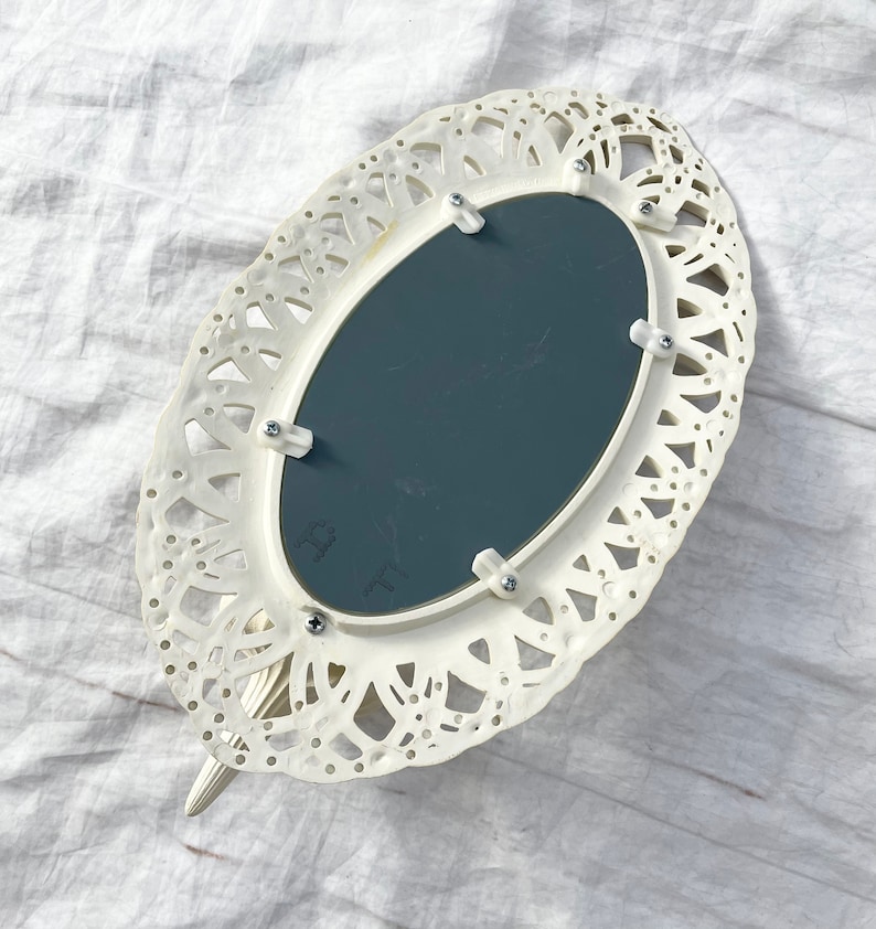 May include: Ivory-colored, oval-shaped hand mirror with a decorative, openwork frame. The mirror has a dark gray reflective surface and is attached with small screws. The handle is a continuation of the frame's design.