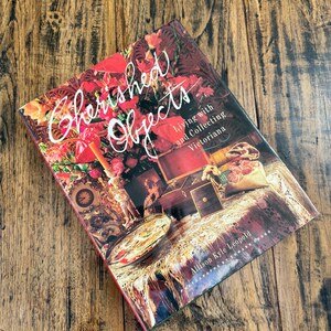 May include: A hardcover book titled Cherished Objects: Living with and Collecting Victoriana by Allison Kyle Leopold. The cover features a dark, floral-themed photograph of antique items, including a clock, a wooden box, and lace.