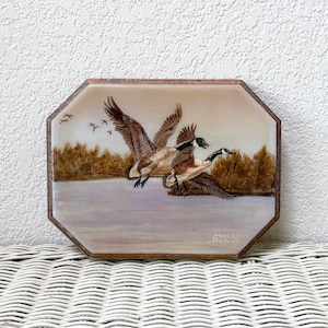 May include: An octagonal decorative plaque featuring a painted landscape with geese in flight over a body of water. The artwork includes trees and a flock of birds in the background. The plaque has a wooden frame and is signed by the artist.