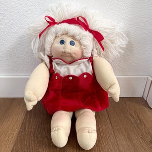May include: A vintage Cabbage Patch Kids doll with a white yarn hairstyle and red ribbon accents. The doll wears a red velvet dress with white trim and buttons. The doll has blue eyes and a soft, plush body.