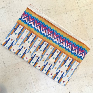 May include: A rectangular, folded textile with a Southwestern-inspired design. The fabric features a cream-colored base with vertical stripes in blue, orange, and yellow. A border with a geometric pattern in red, blue, and gold accents the top edge.
