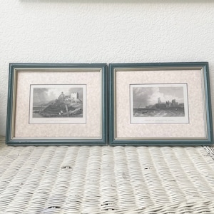 May include: Two framed vintage black and white landscape prints. Each print features a castle or fortress scene. The frames are a muted teal color with a gold inner border. The prints are displayed on a white wicker table.
