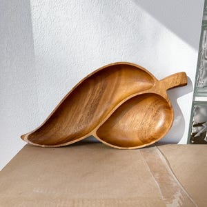 Può includere: Un vassoio da portata in legno a forma di foglia con due scomparti e un manico. Il vassoio è realizzato in legno marrone chiaro e presenta un motivo a venature naturali. Progettato per servire snack o antipasti.