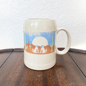 Puede incluir: Taza de cerámica color crema con un diseño de paisaje desértico. El diseño presenta un gran sol blanco, cielo azul, desierto naranja y cactus verdes. Dos siluetas de animales blancos están en primer plano. La taza tiene un asa grande y está sobre una superficie de madera.