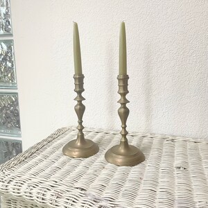 May include: Two antique brass candlesticks with green taper candles. The candlesticks have a detailed design and are placed on a white wicker surface. The candles are a light green color.