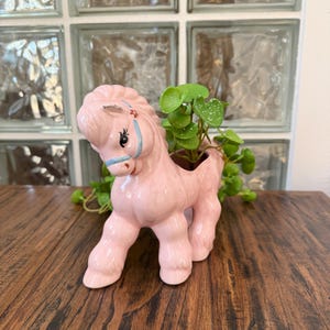 May include: A pink ceramic horse planter with a small green plant inside. The horse has a blue bridle and a decorative mane. The planter is on a wooden surface.