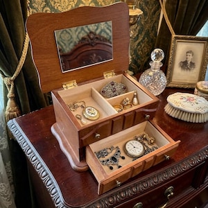 Op de afbeelding: Een houten sieradendoos met een spiegel in het deksel, open om compartimenten met sieraden te onthullen. De doos staat op een donker houten oppervlak, met een lade uitgetrokken, met meer sieraden. De doos is bruin met gouden scharnieren.
