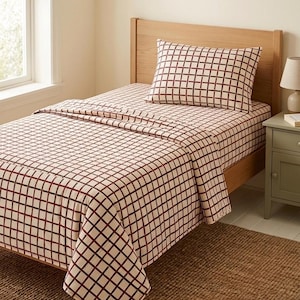 May include: A bed with a cream-colored sheet set featuring a dark red grid pattern. The set includes a fitted sheet, a flat sheet, and a pillowcase. The bed frame is made of light-colored wood.