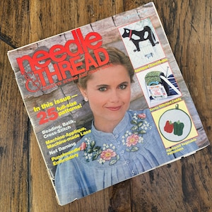 May include: A vintage copy of "Needle & Thread" magazine. The cover features a woman in a blue blouse with floral embroidery. Text includes "25 full-size patterns" and various needlecraft techniques. The magazine is on a wooden surface.