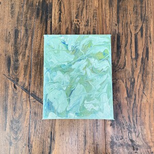 May include: A small, rectangular canvas with an abstract painting. The artwork features a blend of light green, blue, and white hues, with a fluid, marbled effect. Thin white lines are painted across the surface, adding texture.