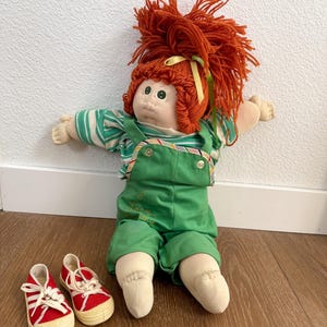 May include: A vintage doll with red yarn hair, green eyes, and a green and white striped shirt. The doll wears green overalls and has a pair of red canvas sneakers with white laces. The doll is a classic toy.