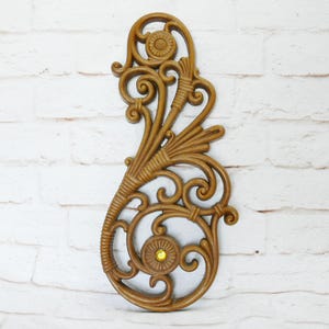 May include: Ornate, brown wall decor with a scrolling design and a yellow gem in the center. The decorative piece features floral and leaf-like elements, creating a vintage aesthetic. The wall hanging is against a white brick background.