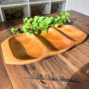 May include: A wooden serving tray with three compartments. The tray is made of light-colored wood and has a smooth finish. A sprig of green leaves rests on the tray. The tray is on a dark wood surface.
