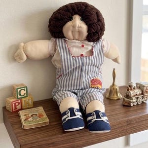 Op de afbeelding: Een stoffen pop met donkerbruin wollen haar, gekleed in een wit overhemd met kraag en een blauw-wit gestreepte tuinbroek met een kleine patch. De pop draagt donkerblauwe stoffen schoenen met witte veters en zit rechtop met gespreide armen.