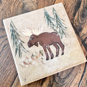 May include: A square stone tile featuring a hand-painted brown moose with large antlers standing in a forest scene. The design includes two green pine trees on either side of the moose and white painted accents representing snow on the ground.
