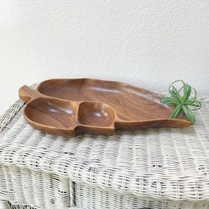 Puede incluir: Una bandeja de madera para servir con diseño de hoja. La bandeja está dividida en tres secciones: un compartimento largo y ovalado y dos compartimentos cuadrados más pequeños. La madera tiene un tono marrón cálido. Una pequeña planta verde está a la derecha.