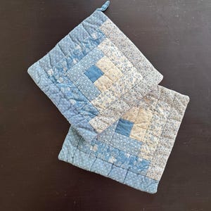 Op de afbeelding: Twee vierkante, gewatteerde pannenlappen in blauw- en wittinten. De pannenlappen hebben een blokhutontwerp met een centraal vierkant en omliggende rechthoekige stukken. Eén pannenlap heeft een lus om op te hangen.