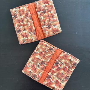 May include: Two quilted fabric book covers. The covers feature a fall-themed pattern with trees in shades of orange, red, and brown. The spine is a solid orange fabric. The covers are placed on a dark surface.