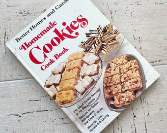 Könnte beinhalten: Ein Kochbuch mit dem Titel "Better Homes and Gardens Homemade Cookies Cook Book" mit Bildern von verschiedenen Keksen. Das Cover zeigt roten und weißen Text, und das Buch ist geöffnet, um Keksrezepte und Fotos anzuzeigen.