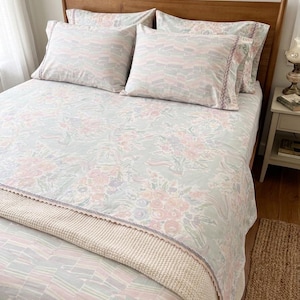 May include: A bed with a wooden frame is dressed with a floral patterned bedspread in pastel shades. Two pillows with a geometric design are placed on top. A cream blanket with a decorative edge is folded at the foot of the bed.