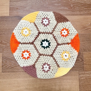 May include: A round, crocheted cushion featuring a patchwork design of hexagonal shapes. The cushion showcases a palette of beige, brown, orange, yellow, and green. The center of each hexagon has a floral design.