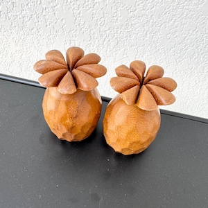 Pode incluir: Dois objetos de madeira artesanais em forma de abacaxi. Cada um tem um corpo texturizado castanho claro e um topo em forma de flor feito de peças sobrepostas em forma de pétala. Os objetos estão em uma superfície preta contra uma parede branca.