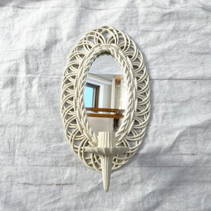 May include: Off-white oval wall mirror with a woven design frame and a candle holder. The mirror is approximately 12 inches tall and 8 inches wide. The candle holder is positioned below the mirror.