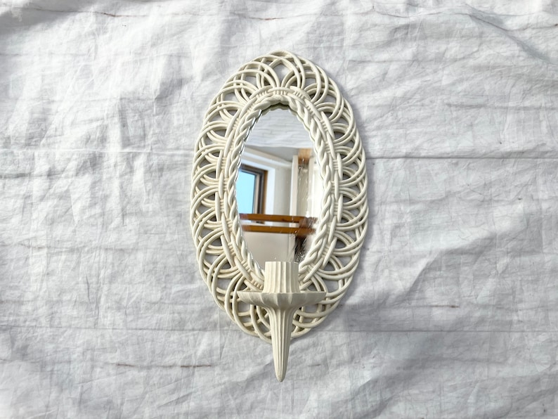 May include: Off-white oval wall mirror with a woven design frame and a candle holder. The mirror is approximately 12 inches tall and 8 inches wide. The candle holder is positioned below the mirror.