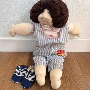 Peut inclure: Une poupée vintage avec une coiffure en fil marron, des yeux verts et un corps en tissu doux. La poupée porte une salopette rayée bleue et blanche avec une bordure rouge et des chaussures bleues assorties.