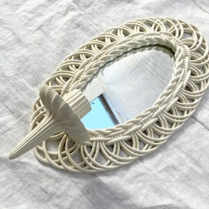 May include: An oval-shaped, off-white woven mirror with a decorative candle holder attached. The mirror has a woven border and a small, decorative candle holder on the side. The candle holder is also off-white and has a textured design.