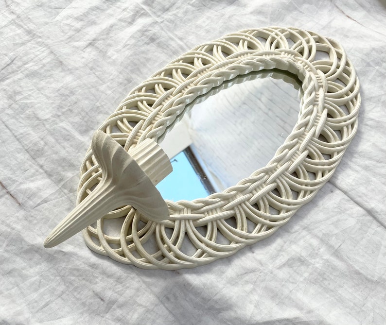May include: An oval-shaped, off-white woven mirror with a decorative candle holder attached. The mirror has a woven border and a small, decorative candle holder on the side. The candle holder is also off-white and has a textured design.