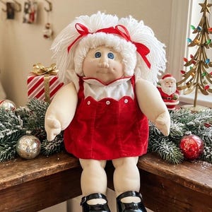 May include: A soft-bodied doll with white yarn hair tied with red ribbons, wearing a red velvet dress with a white collar and scalloped trim, and black patent leather-style shoes. The doll has blue eyes and a light skin tone, sitting upright.