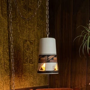 May include: A hanging pendant lamp featuring a white cylindrical shade with a tapered top. The lower section displays a geometric pattern in black and brown with triangular cutouts that emit a warm light, suspended by a long metal chain.