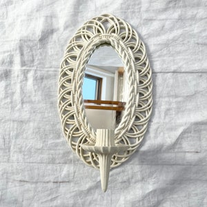 May include: Off-white oval wall mirror with a woven rattan-style frame. The mirror has a built-in candle holder. The mirror is approximately 12 inches tall and 8 inches wide.