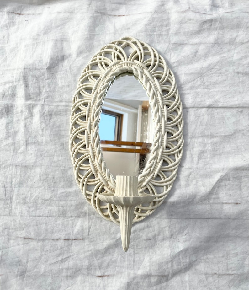 May include: Off-white oval wall mirror with a woven rattan-style frame. The mirror has a built-in candle holder. The mirror is approximately 12 inches tall and 8 inches wide.