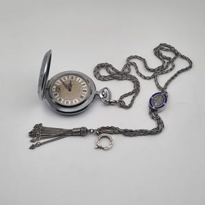 Può includere: Un orologio da tasca argentato con catena e nappa. Il quadrante dell'orologio ha numeri romani e uno sfondo chiaro. La catena ha un elemento decorativo blu e argento.