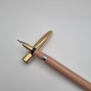 May include: A gold fountain pen with a light brown barrel and silver detailing. The cap is removed, showing the nib. The pen rests on a white surface.