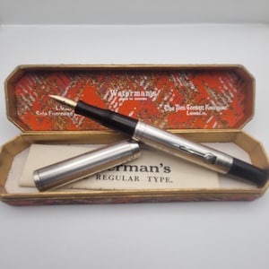 May include: A vintage Waterman's fountain pen with a silver barrel and black accents, displayed in an open, patterned case. The pen's nib is visible, and the case interior features text including "Waterman's" and "Regular Type."