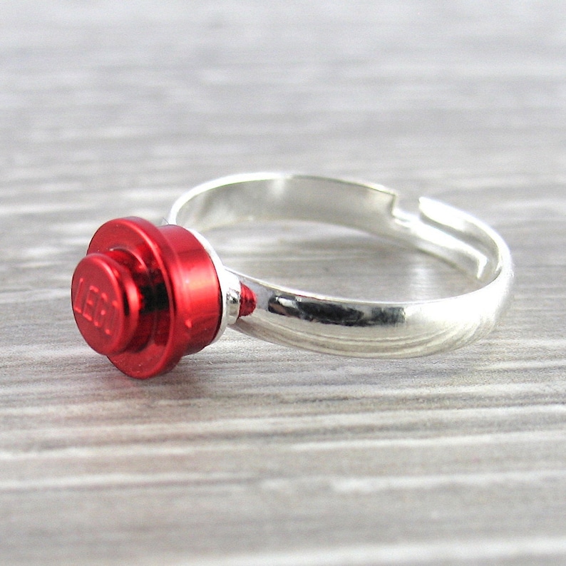 May include: A silver adjustable ring with a red LEGO brick attached to the band. The LEGO brick has the LEGO logo printed on it.