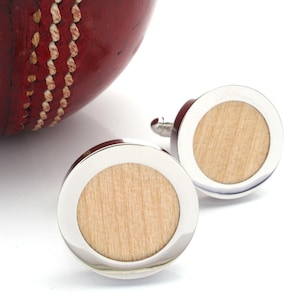 May include: Close-up of a pair of round cufflinks with a light wood center and a silver-colored border. A red cricket ball is partially visible in the background. The cufflinks have a classic design.