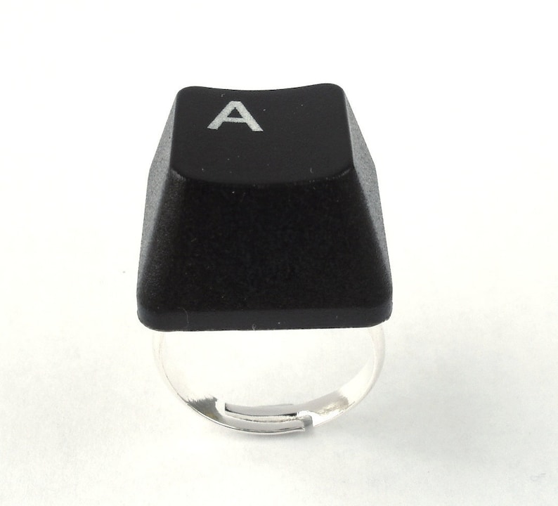 May include: A black keyboard key ring with the letter "A" printed in white on the top. The ring is made of silver metal.