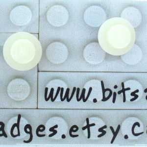 Può includere: Due bottoni di plastica bianca con un centro rialzato, mostrati su uno sfondo a griglia bianca. Il testo "www.bitsandbadges.etsy.com" è stampato sotto la griglia.
